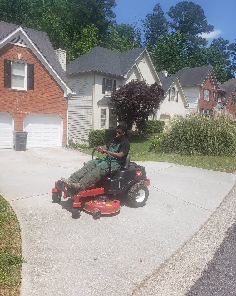 Person riding a red lawn mower on a suburban driveway near houses and trees