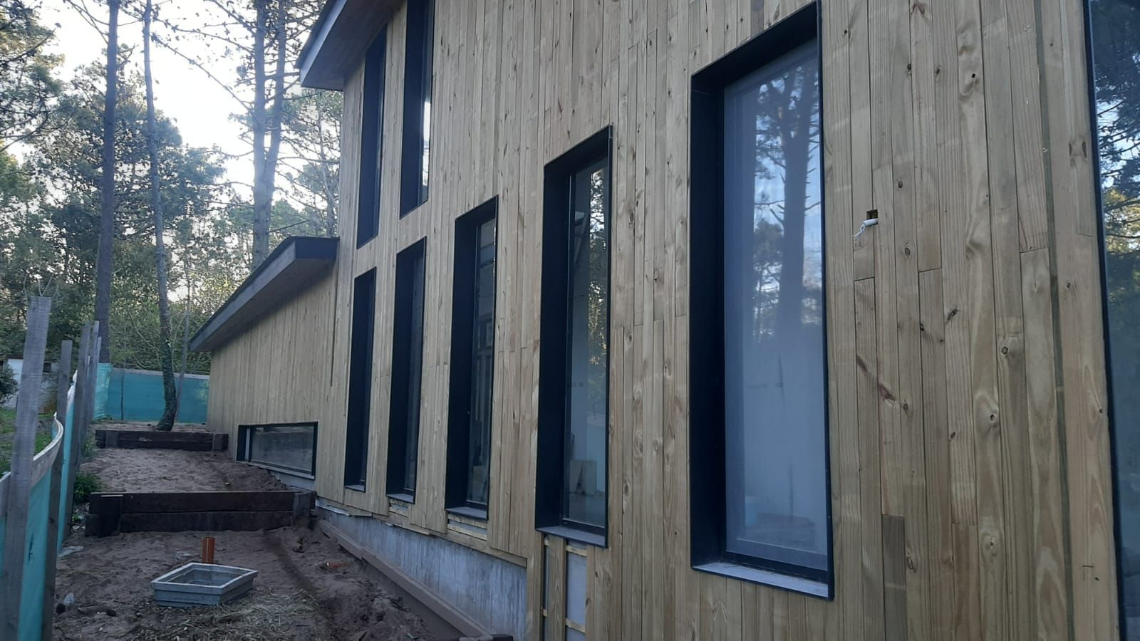 Edificio revestido de madera con ventanas con marco negro; vista exterior en un entorno forestal con un camino de grava.