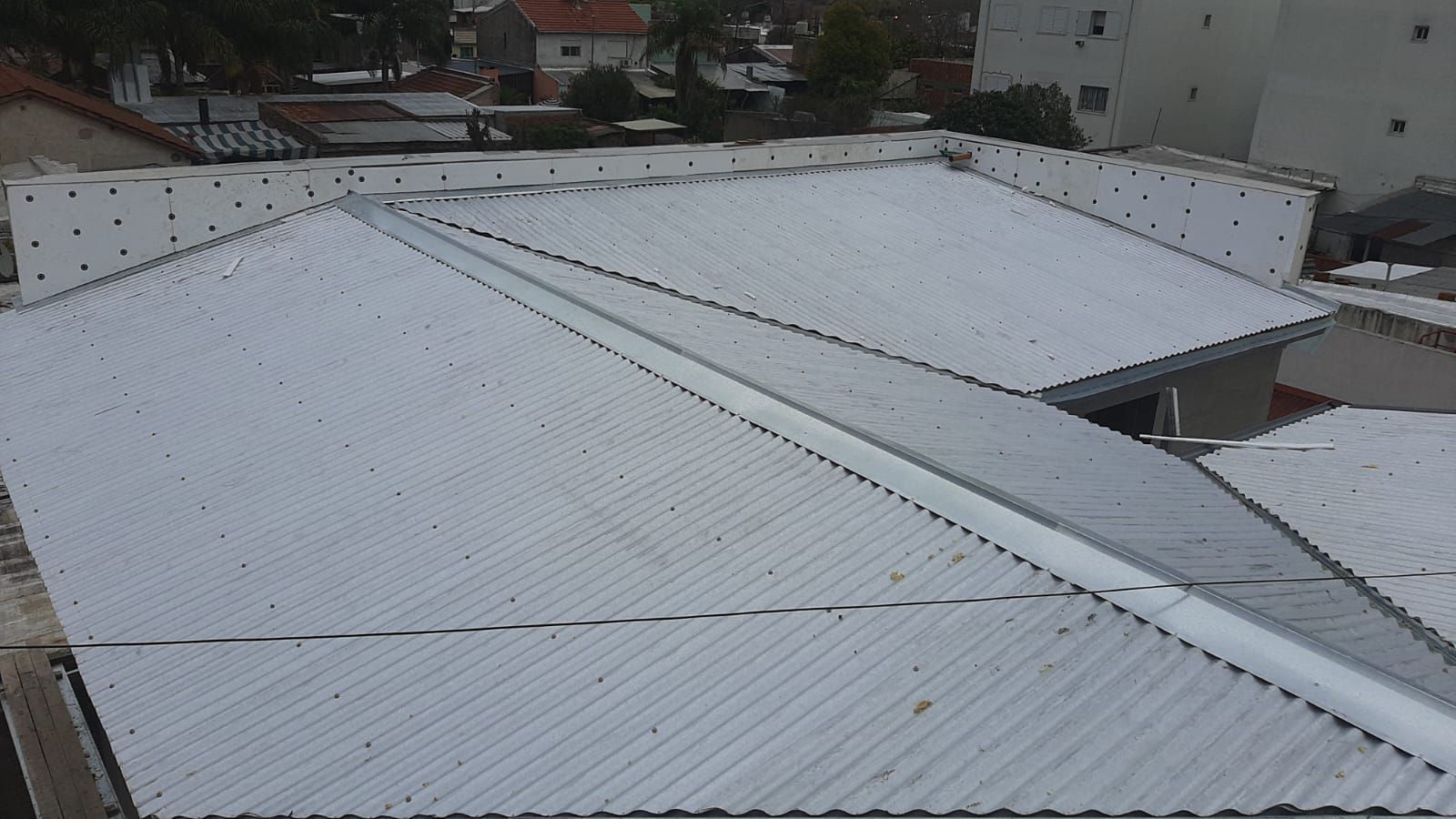 Vista aérea de un techo de metal corrugado plateado con una cumbrera central y edificios circundantes.