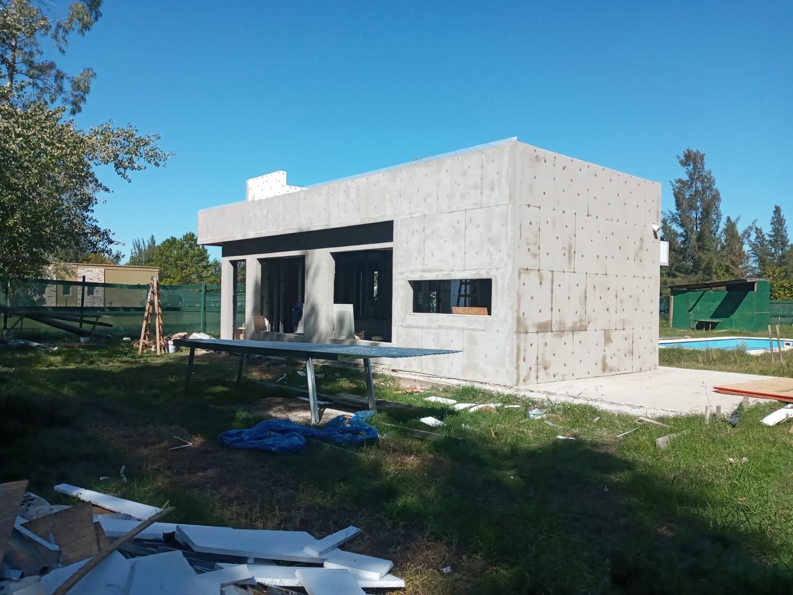 Edificio de hormigón en construcción; vista exterior, cielo despejado, patio con césped, aberturas de ventanas y puertas.