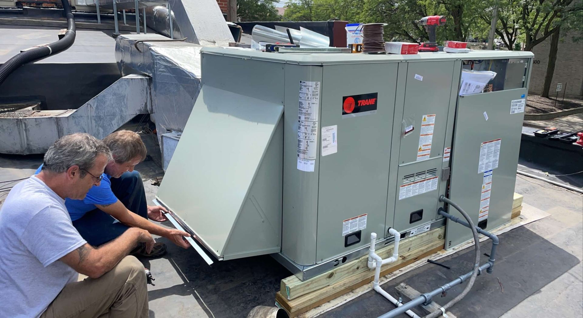 Two men are working on an air conditioner on top of a roof.