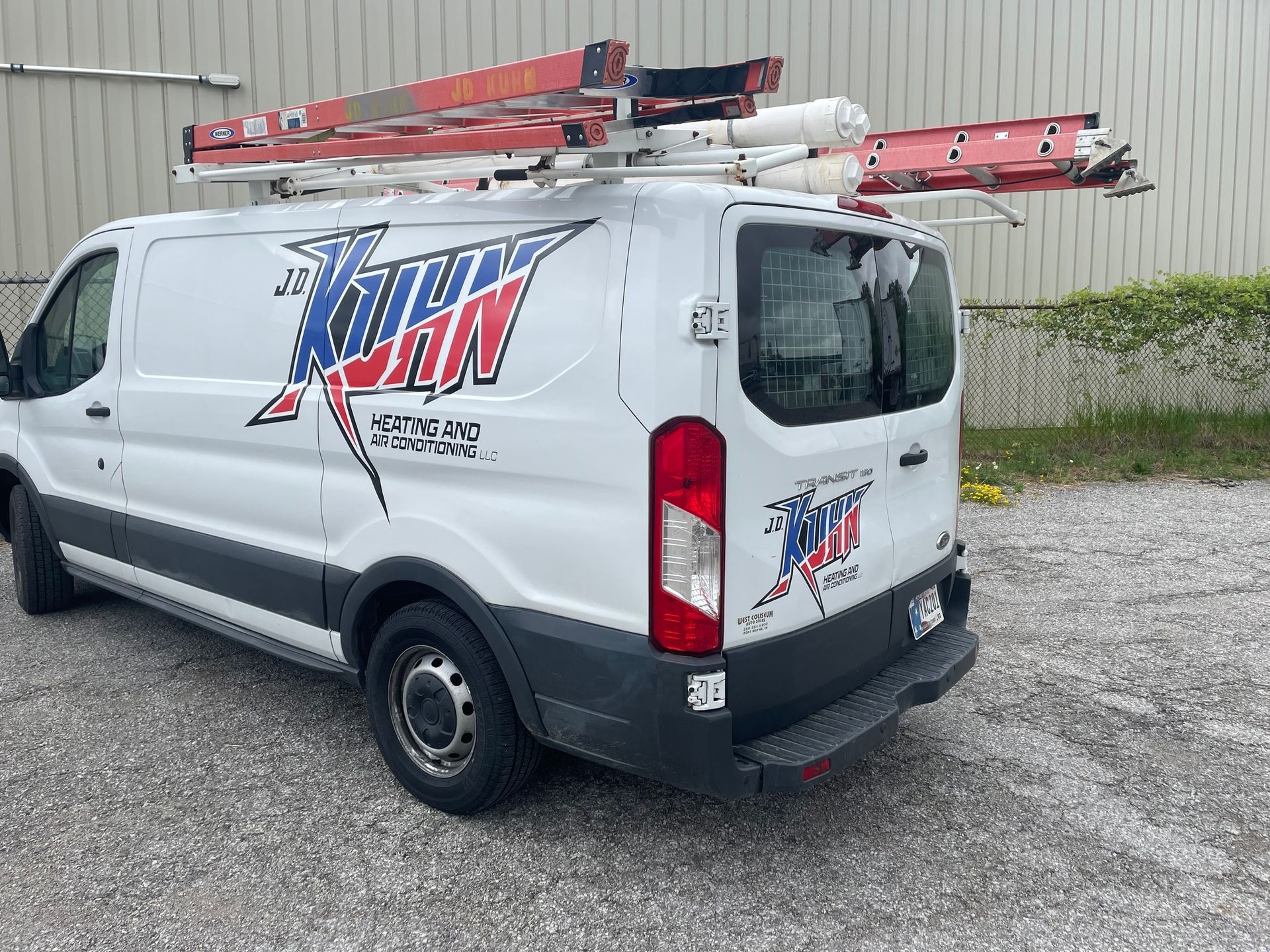 A white van with a ladder on top of it is parked in a gravel lot.