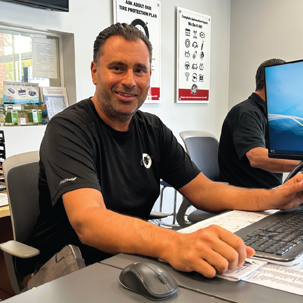 Man in black shirt smiling at a desk, another person blurred in background at computer.