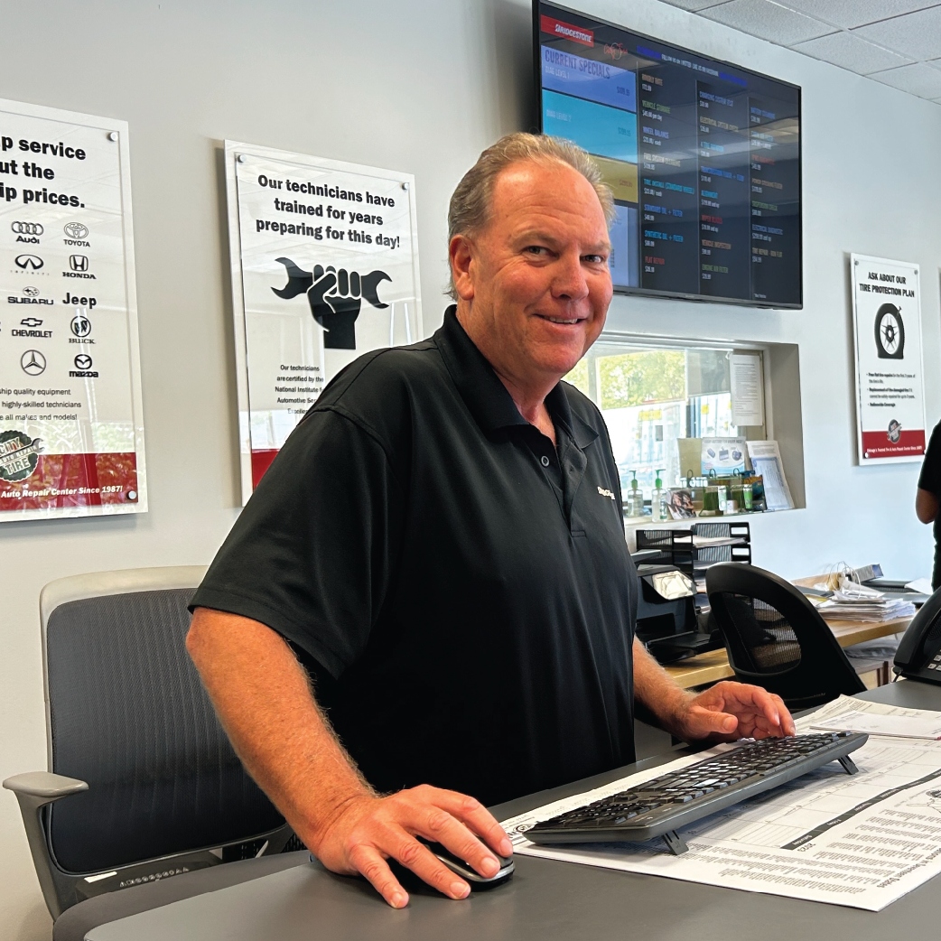Man at a desk in a shop, smiling. Wearing black shirt. Background: posters, computer monitor.
