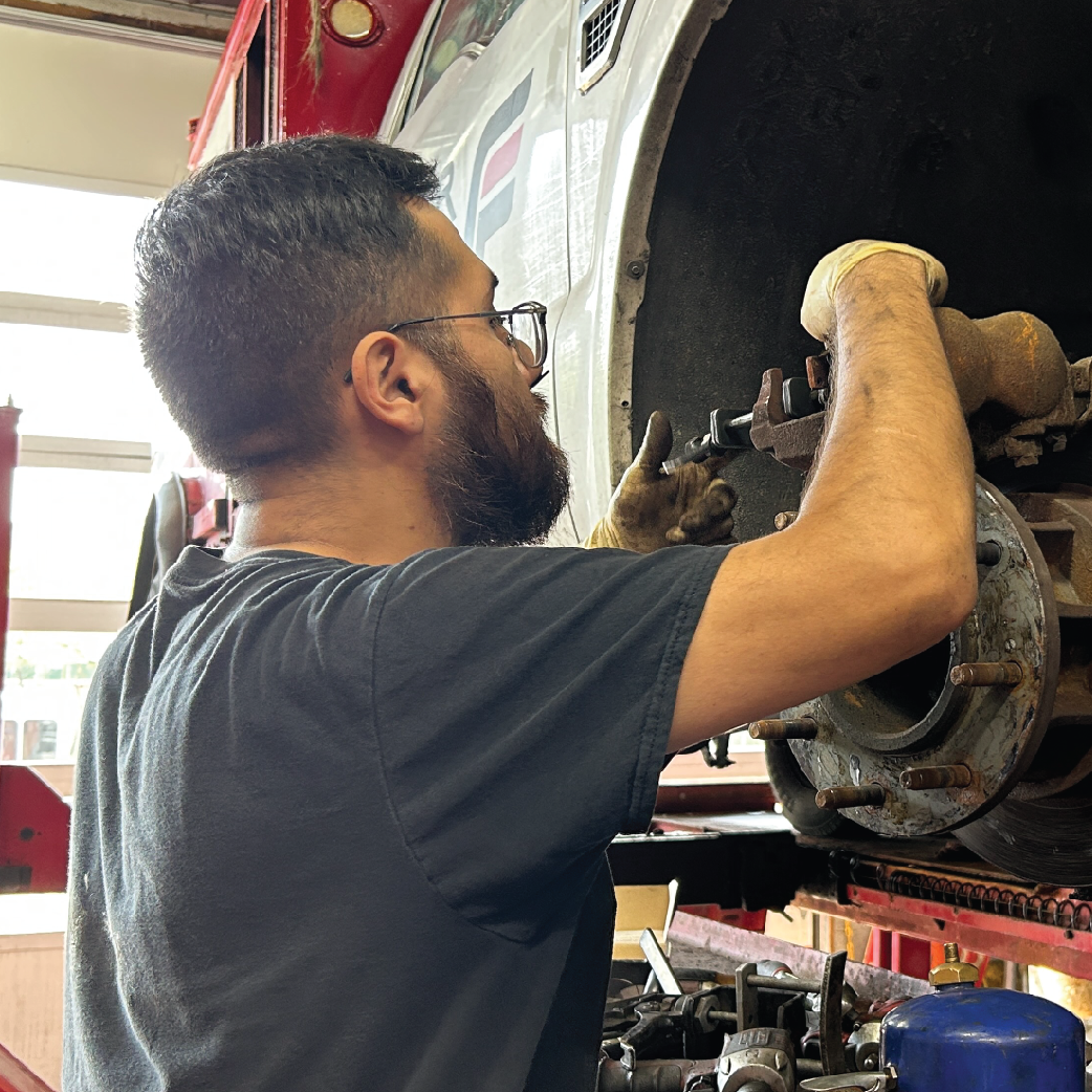 Mechanic with beard and glasses working on a vehicle wheel in a shop, wearing gloves.
