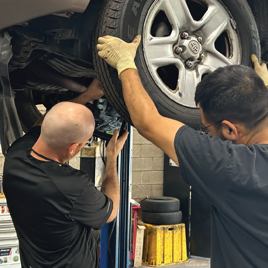 Two mechanics replacing a car tire in a garage; one is holding the tire.