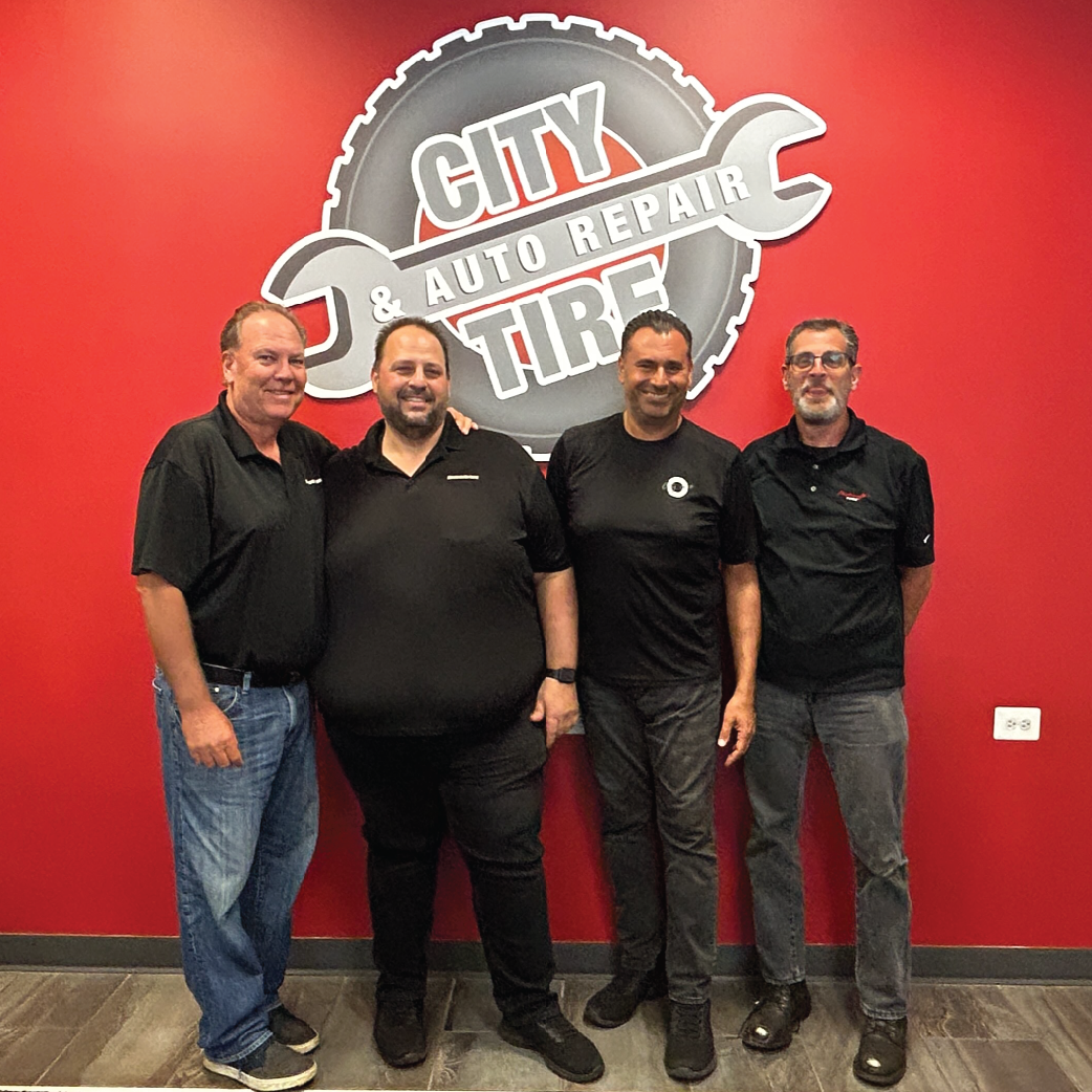 Four men standing in front of a red wall with the logo of a tire and auto repair shop.