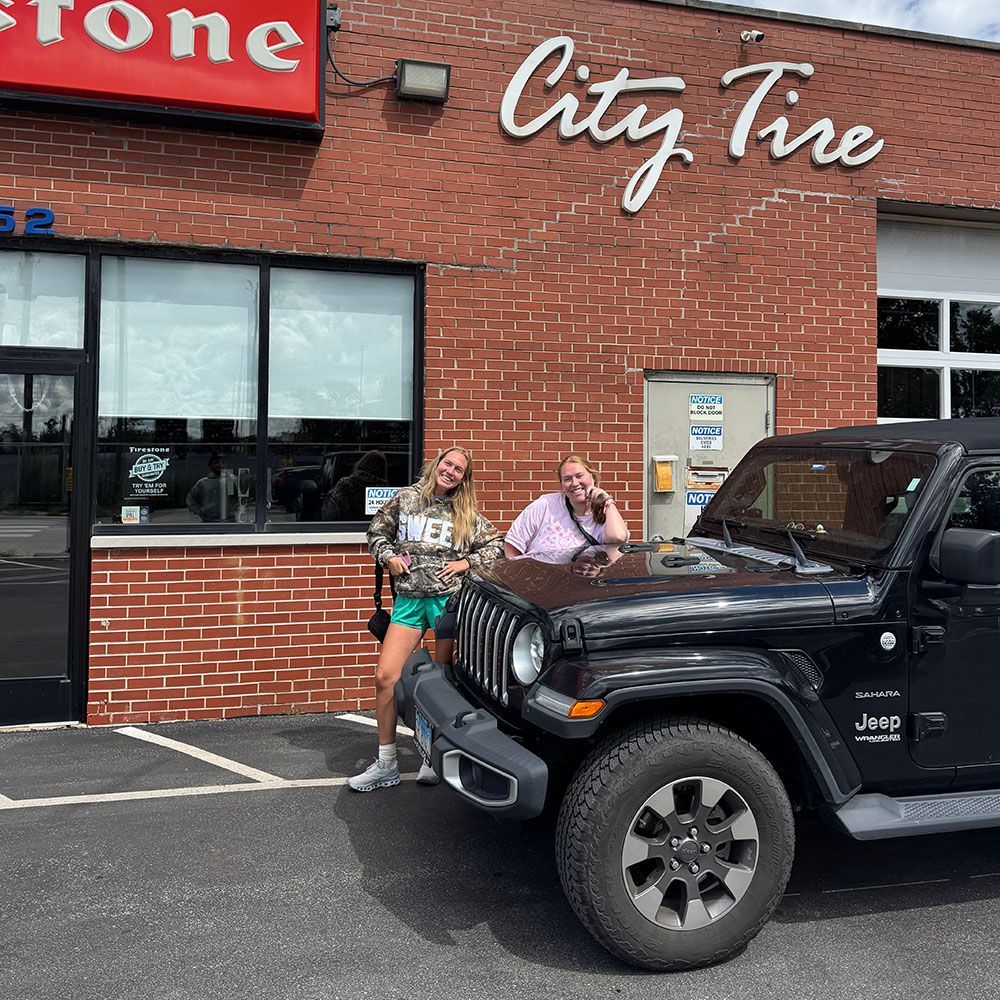 Two women pose with a black Jeep in front of a City Tire shop.