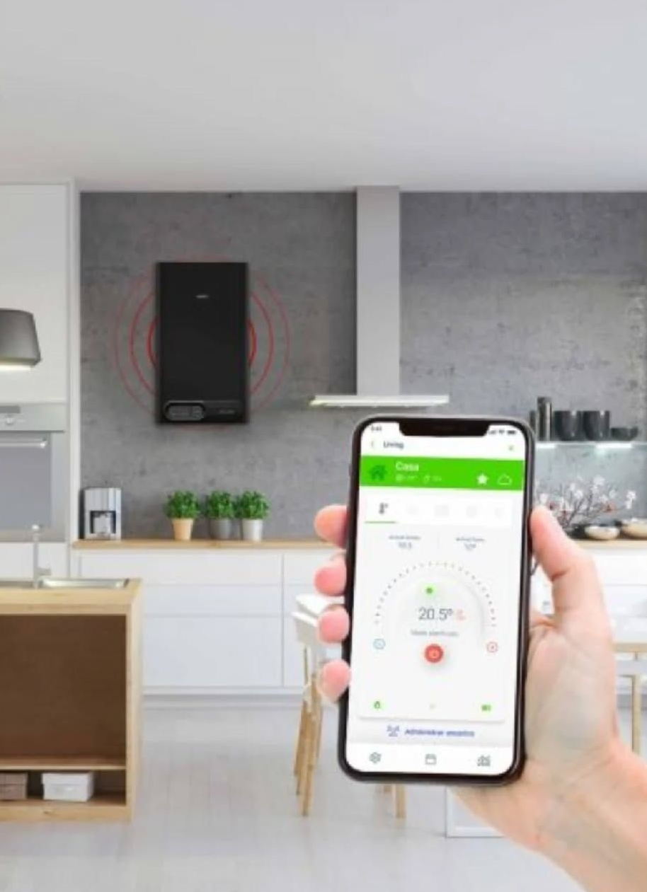 Mano sosteniendo un teléfono mientras se muestra una aplicación en un dispositivo inteligente. Al fondo se observa un dispositivo negro montado en la pared. Escenario de cocina.