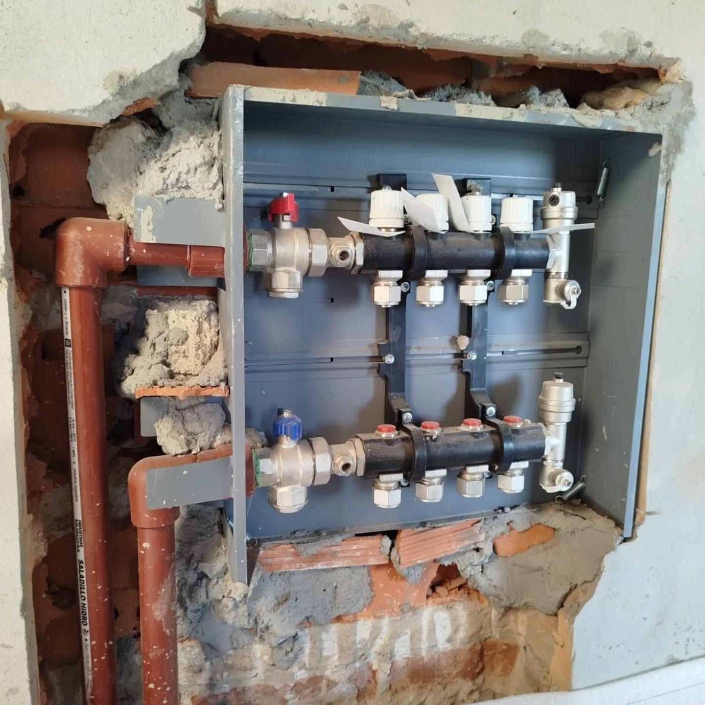 Colector de fontanería con válvulas y tuberías rojas/azules en una cavidad de pared, tonos marrones y grises.