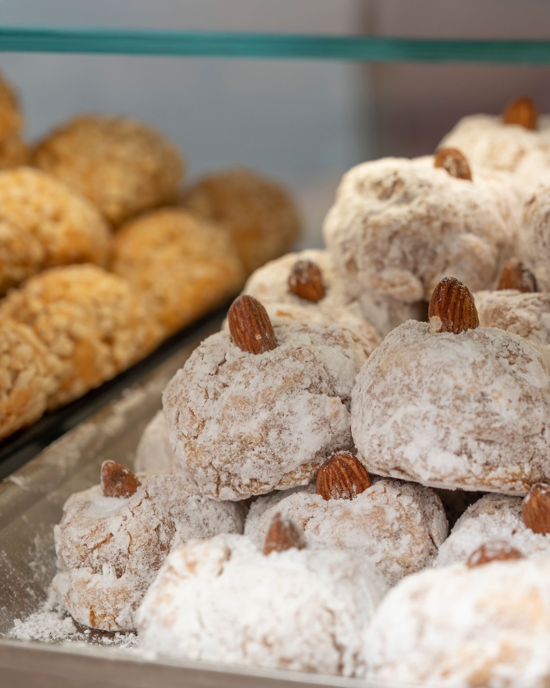 Primo piano di biscotti alle mandorle, alcuni ricoperti di polvere, altri con mandorle, impilati su un vassoio, in una vetrina da pasticceria.