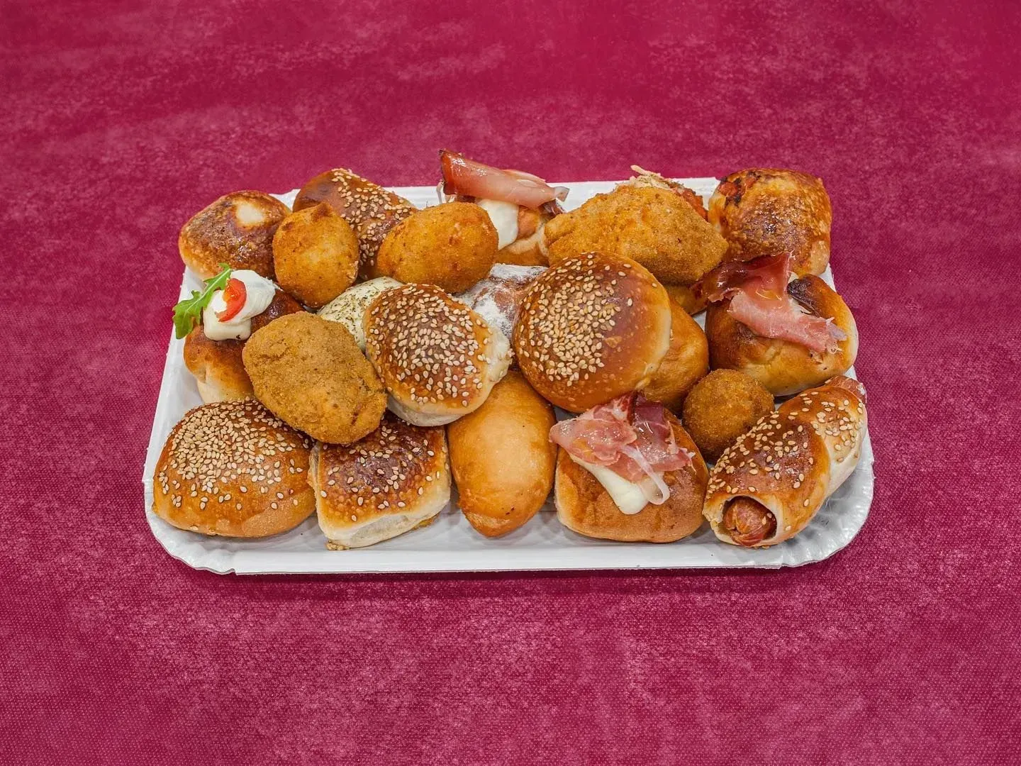 Vassoio di pasticcini salati assortiti su una superficie rossa; alcuni con semi di sesamo, carne o formaggio.