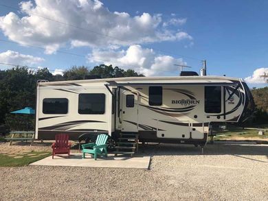 A rv is parked in a gravel lot with chairs and umbrellas.