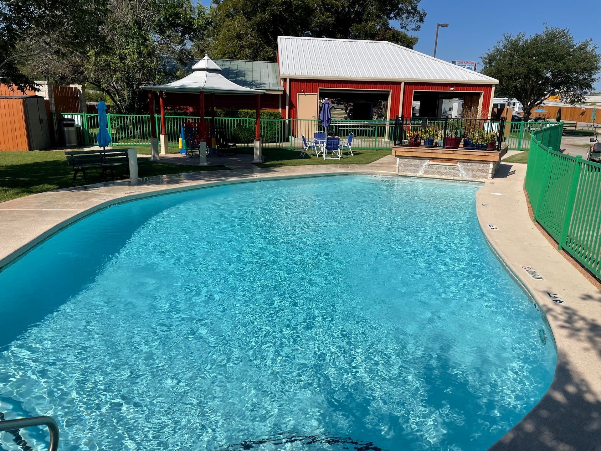 A large swimming pool is surrounded by a green fence and a red building.