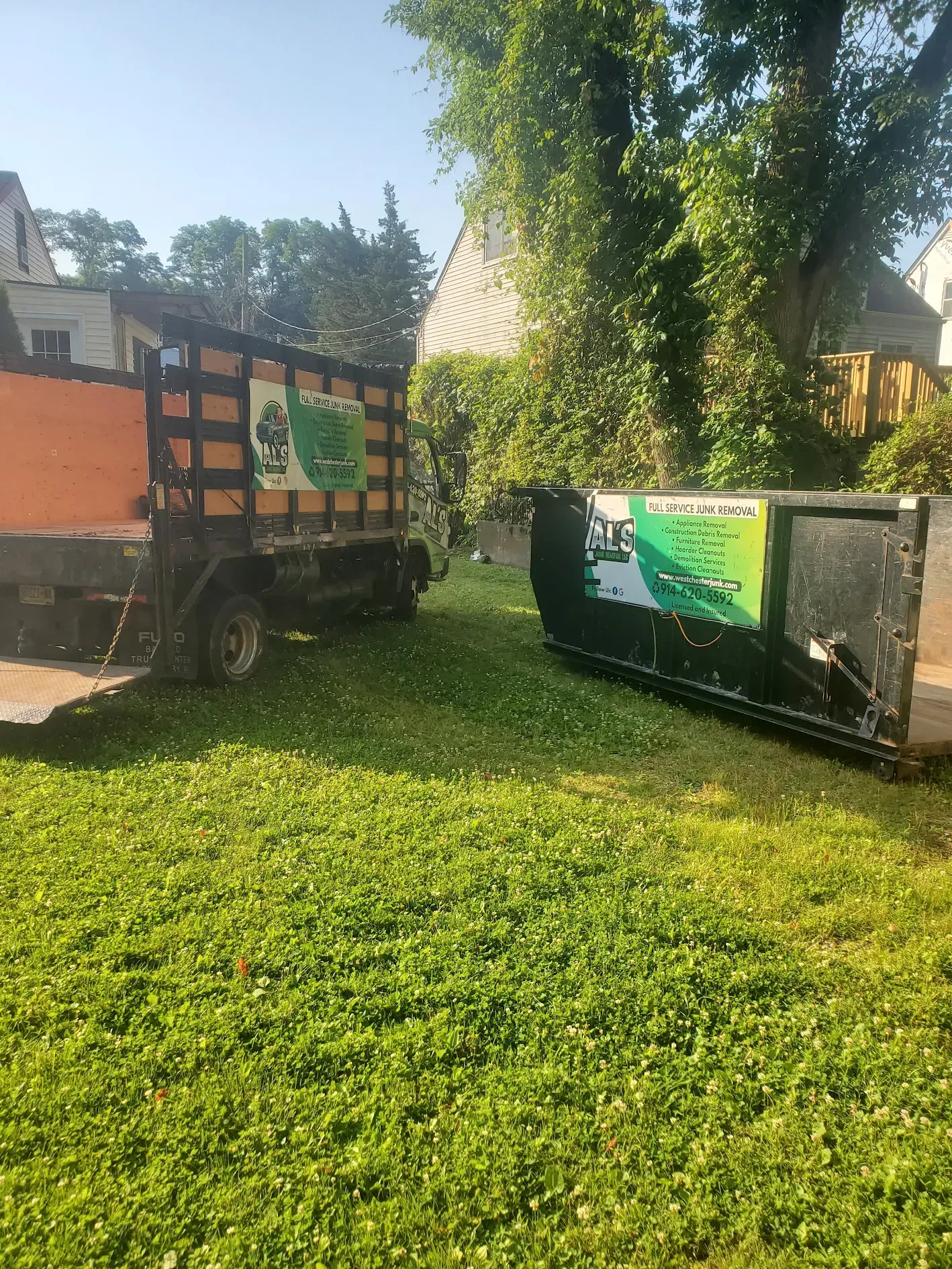 A dump truck and a dumpster are parked in a grassy yard.