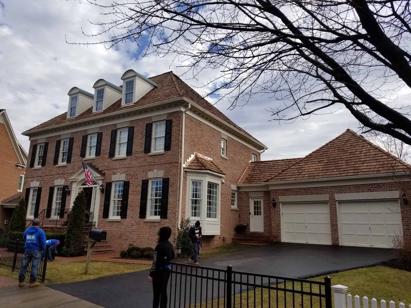 Brick house with a garage, black shutters, and a dark driveway. People stand on the lawn.