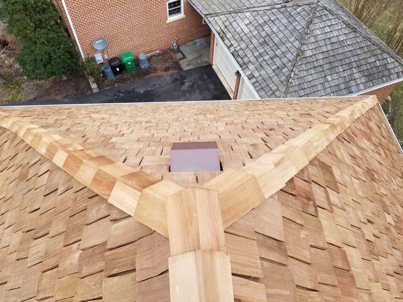 Overhead view of a cedar shake roof with intersecting ridges, a small chimney, and portions of surrounding buildings.