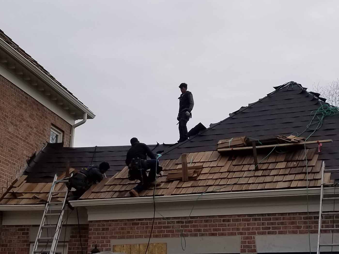 Roofers replacing shingles on a brick building's roof under an overcast sky.