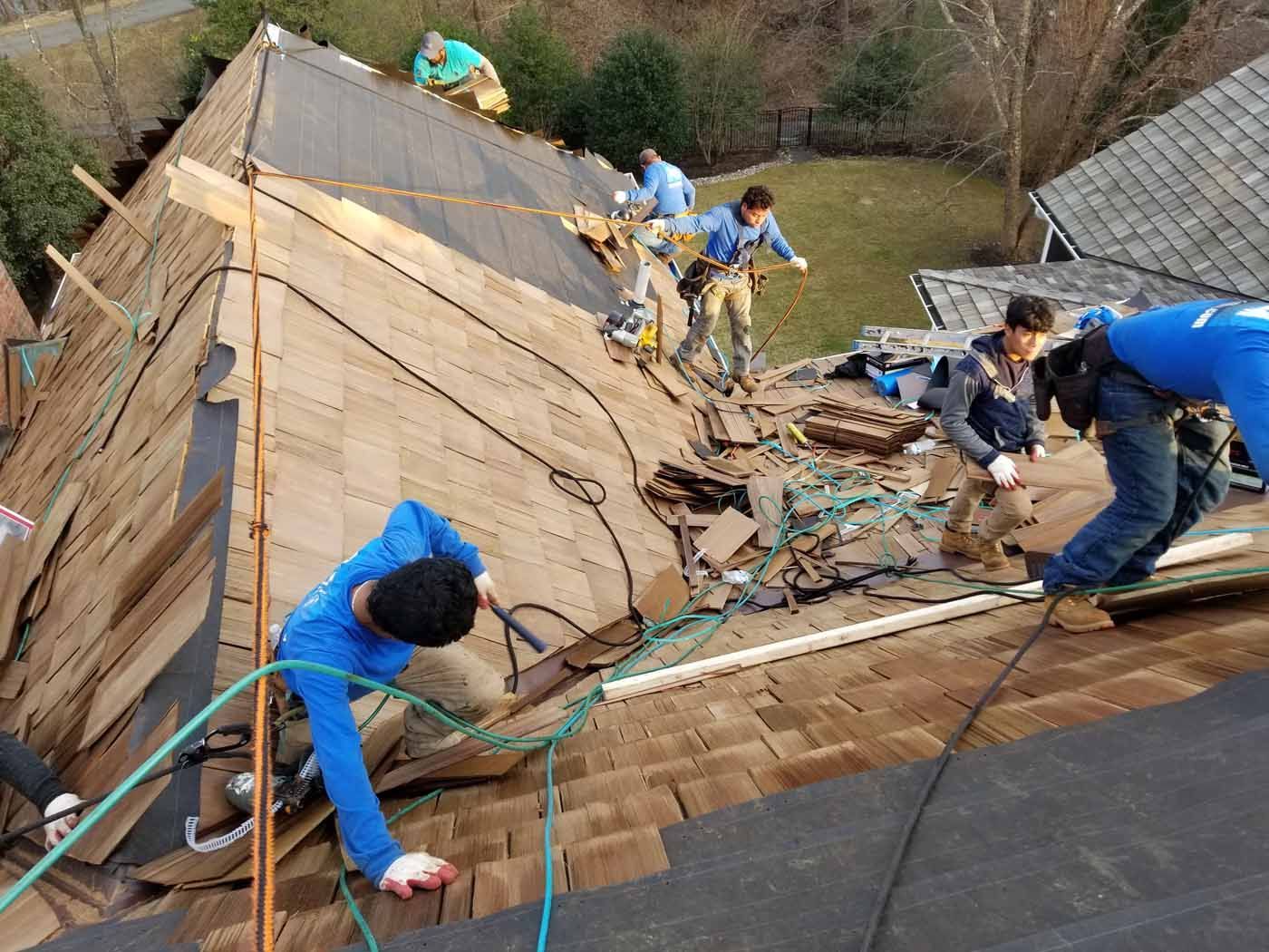Roofers removing old shingles from a house roof. Blue and green safety lines are attached. Brown shingles, sunny day.