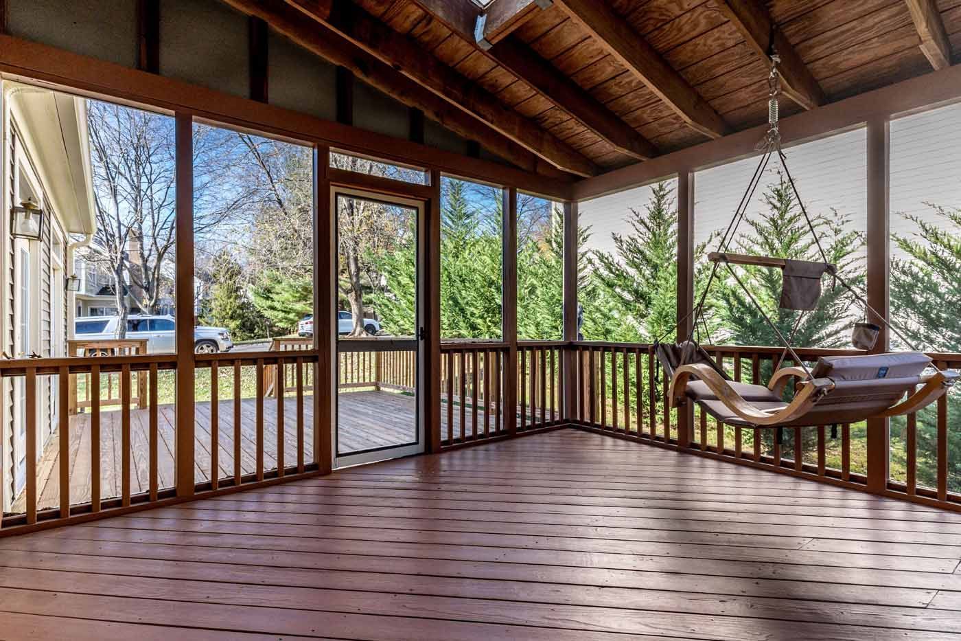 Screened porch with wooden floor, railing, and hanging swing, looking out to trees and a clear sky.