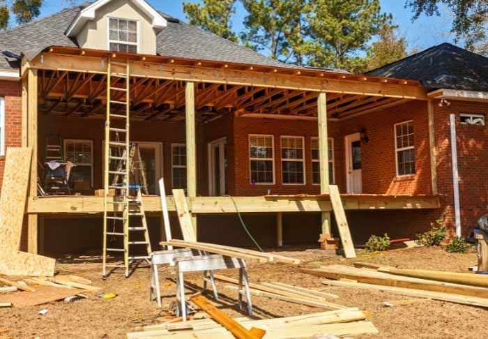 Exterior view of a deck under construction attached to a red brick house; wood frame and lumber visible.