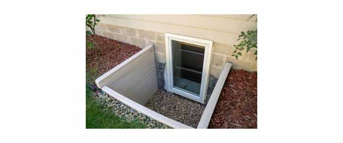 Basement window well with window, surrounded by gravel and mulch. Exterior of a house with grass.
