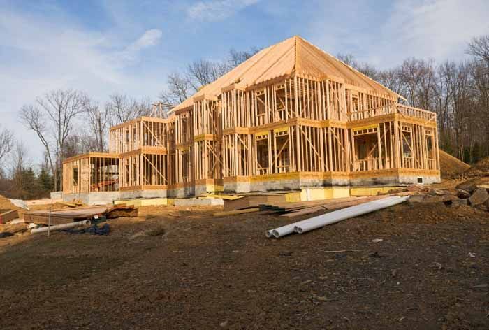 House under construction, wooden frame visible. On a dirt lot, surrounded by bare trees under a blue sky.