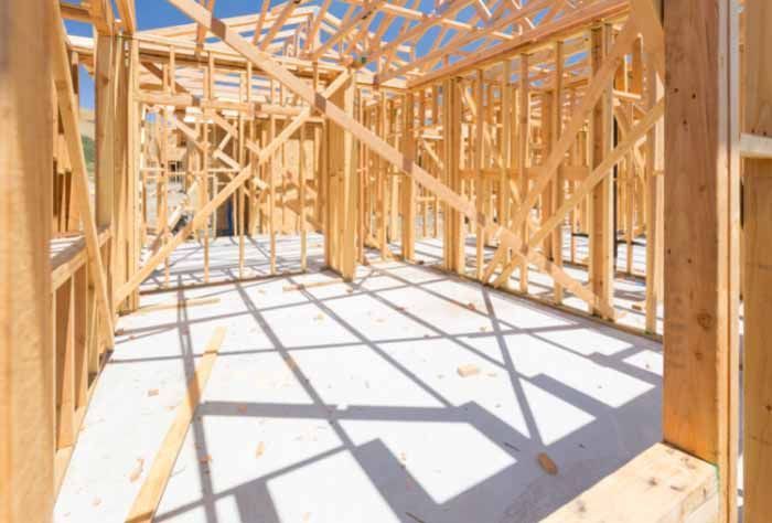 Wooden framework of a building under construction, with a concrete floor and sunlight casting shadows.