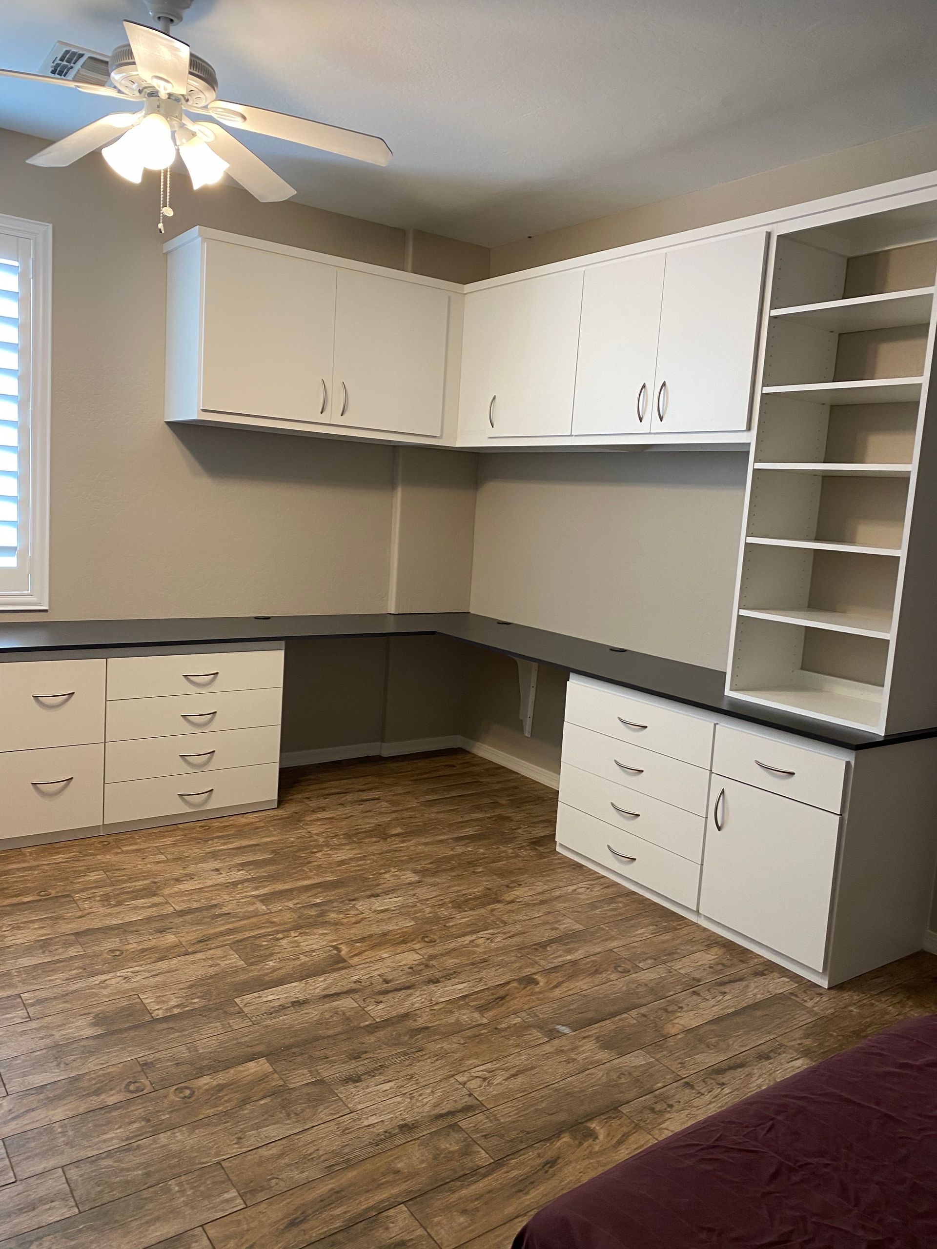 There is a ceiling fan in the home office with a lot of cabinets and drawers.