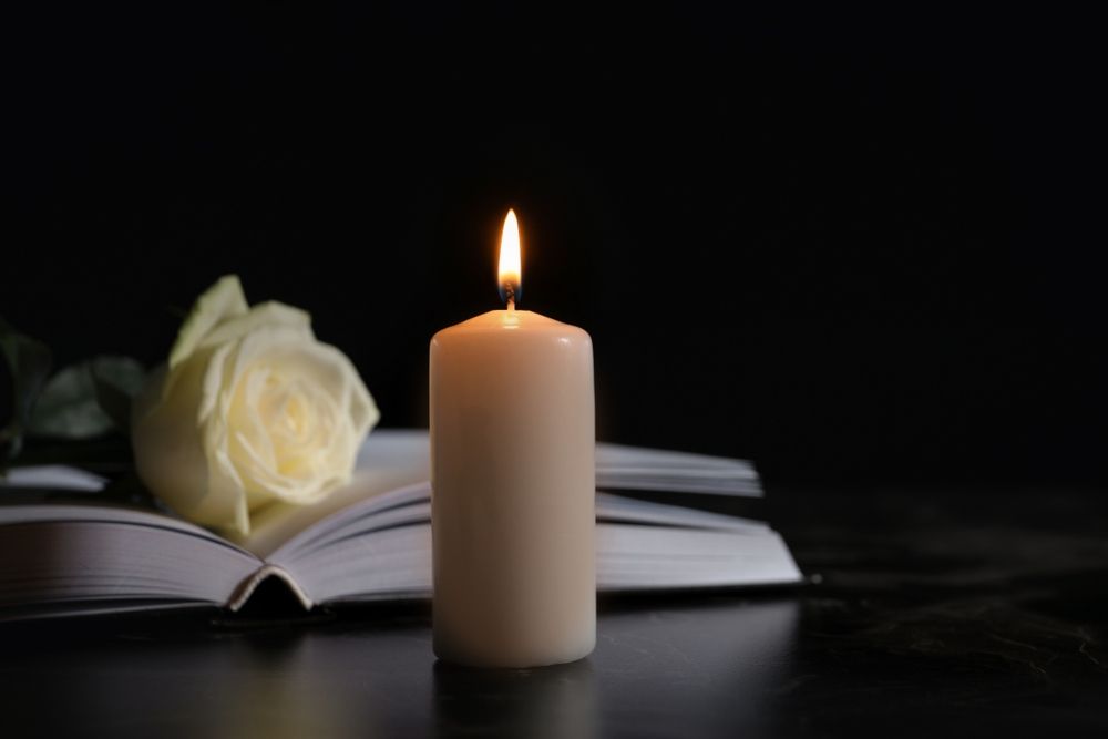 A Lit Candle is Sitting on Top of an Open Book Next to a White Rose — Becker Family Funerals In Taree, NSW