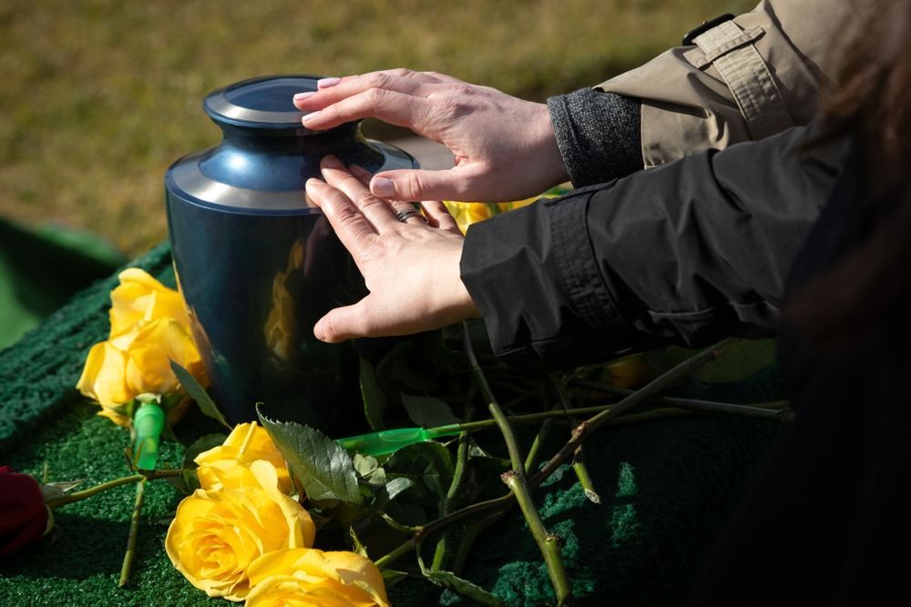 A Person is Touching an Urn With Their Hands Next to Yellow Roses — Becker Family Funerals In Taree, NSW