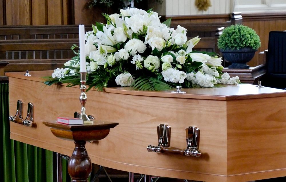 A Wooden Coffin With White Flowers and a Candle in a Church — Becker Family Funerals In Taree, NSW