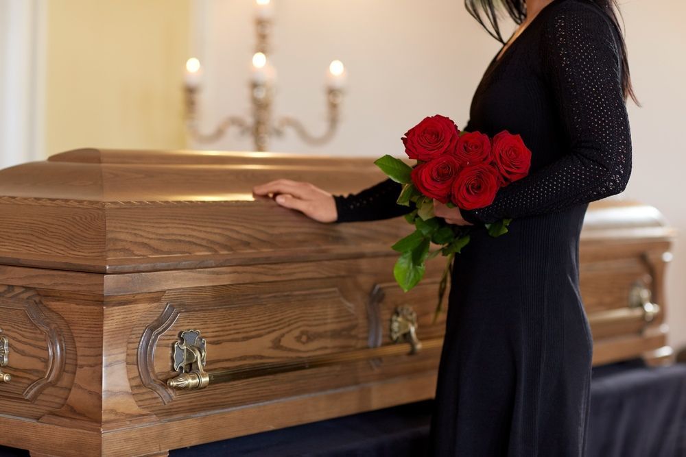 A Woman in a Black Dress is Standing Next to a Coffin Holding Red Roses — Becker Family Funerals In Taree, NSW