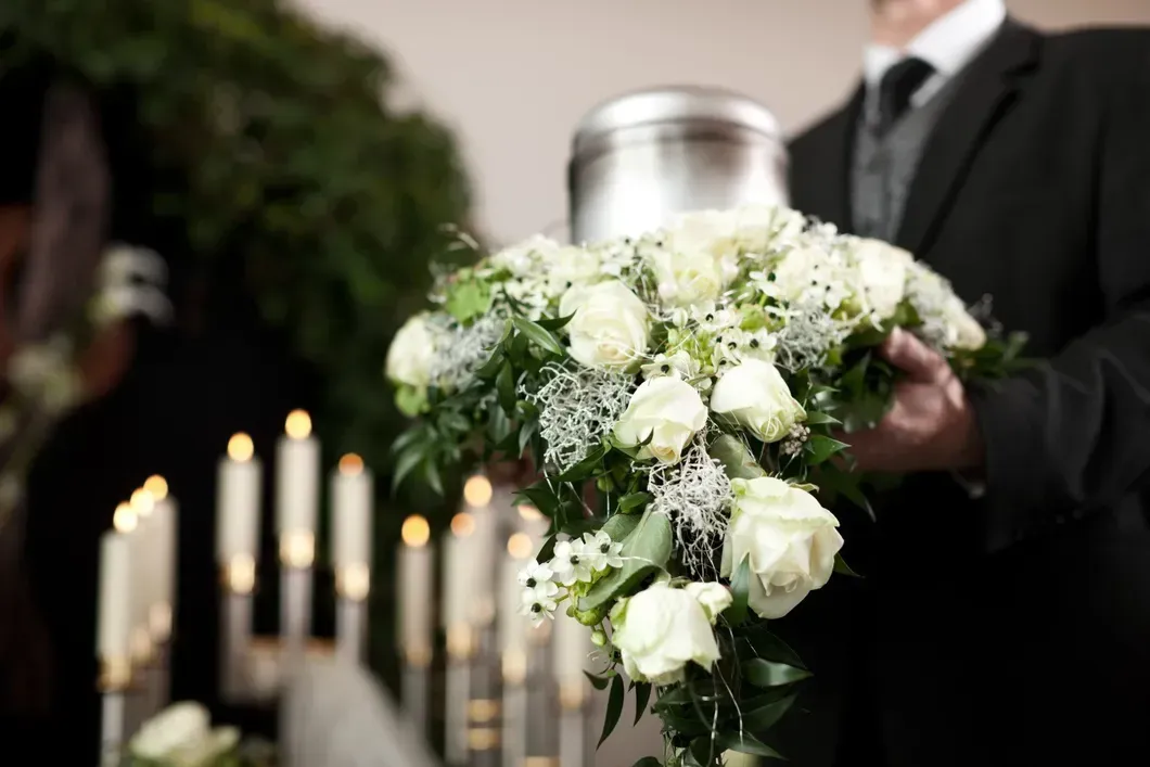 Un mazzo di fiori bianchi per un funerale, accanto a candele accese, tenuto da una persona in abito nero.