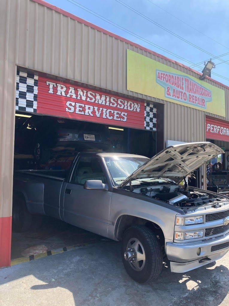 A silver truck with the hood up is parked in front of a transmission service shop.