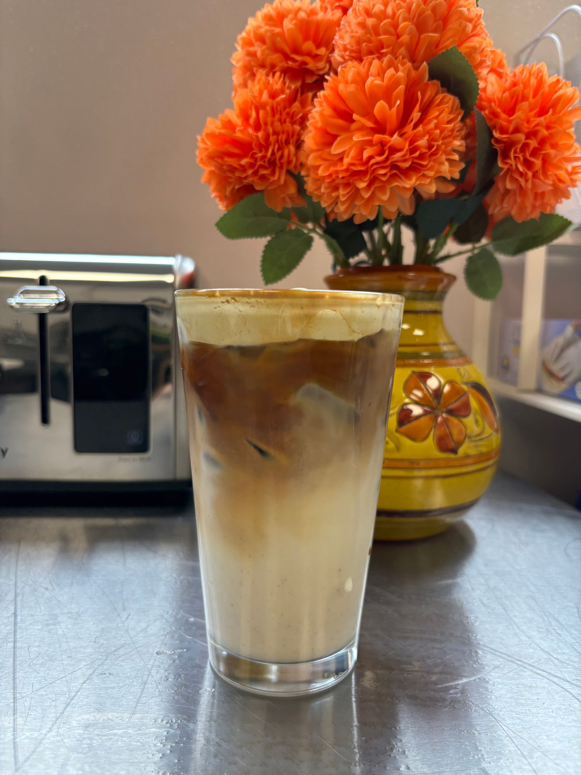 A glass of iced coffee sits on a counter next to a vase of orange flowers