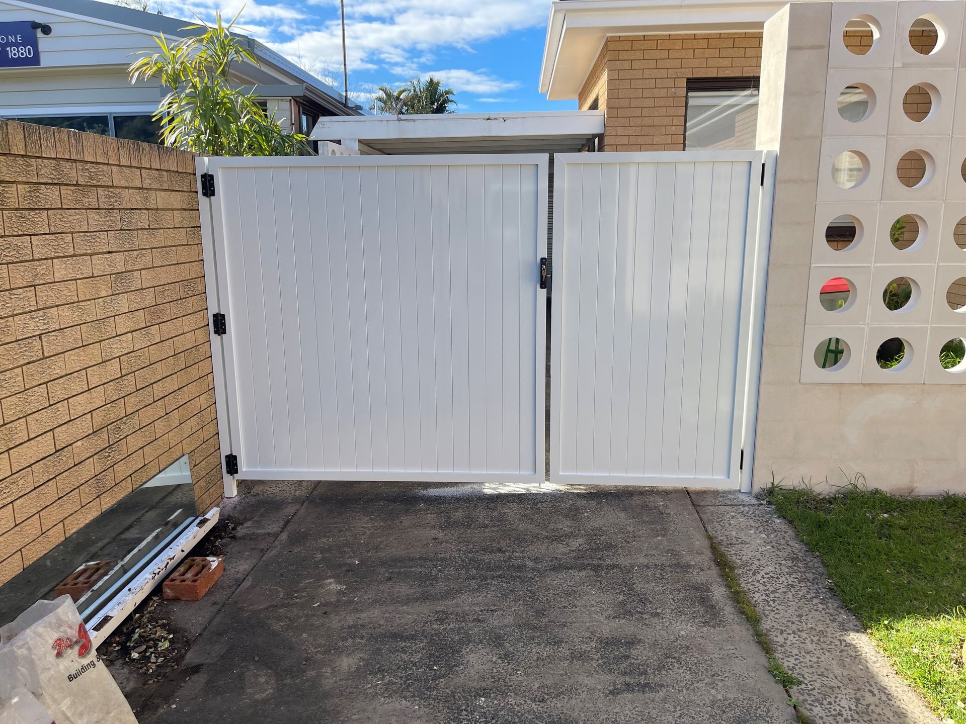 White double driveway gate beside a brick wall and driveway entrance, with a small lawn on the right.