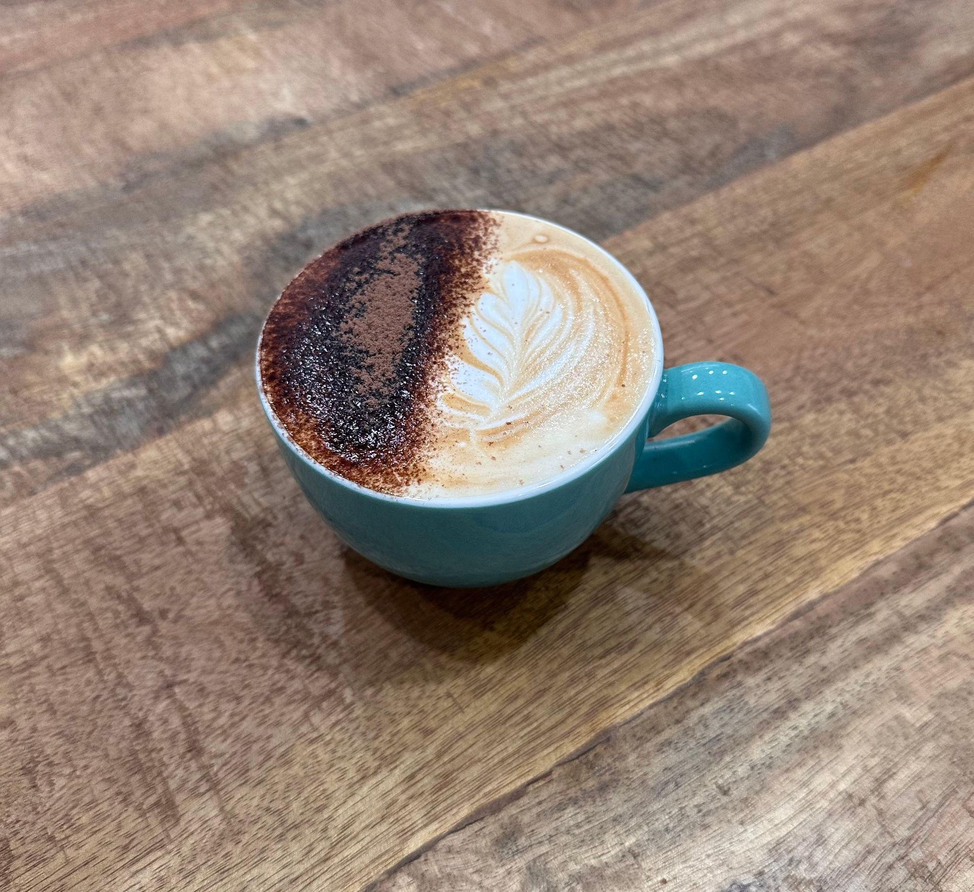 Cappuccino in a teal mug on a wooden table, half with cocoa powder, half with latte art — Labroco Gourmet Coffee Supplier In Forster, NSW