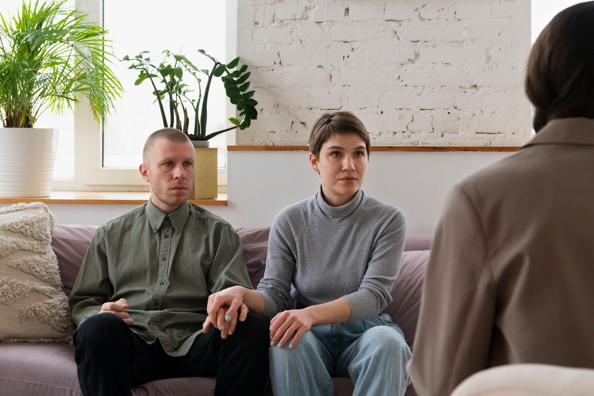 Couple holding hands on couch, facing a person in a neutral-colored jacket, possibly a therapist. Room with plants and window.