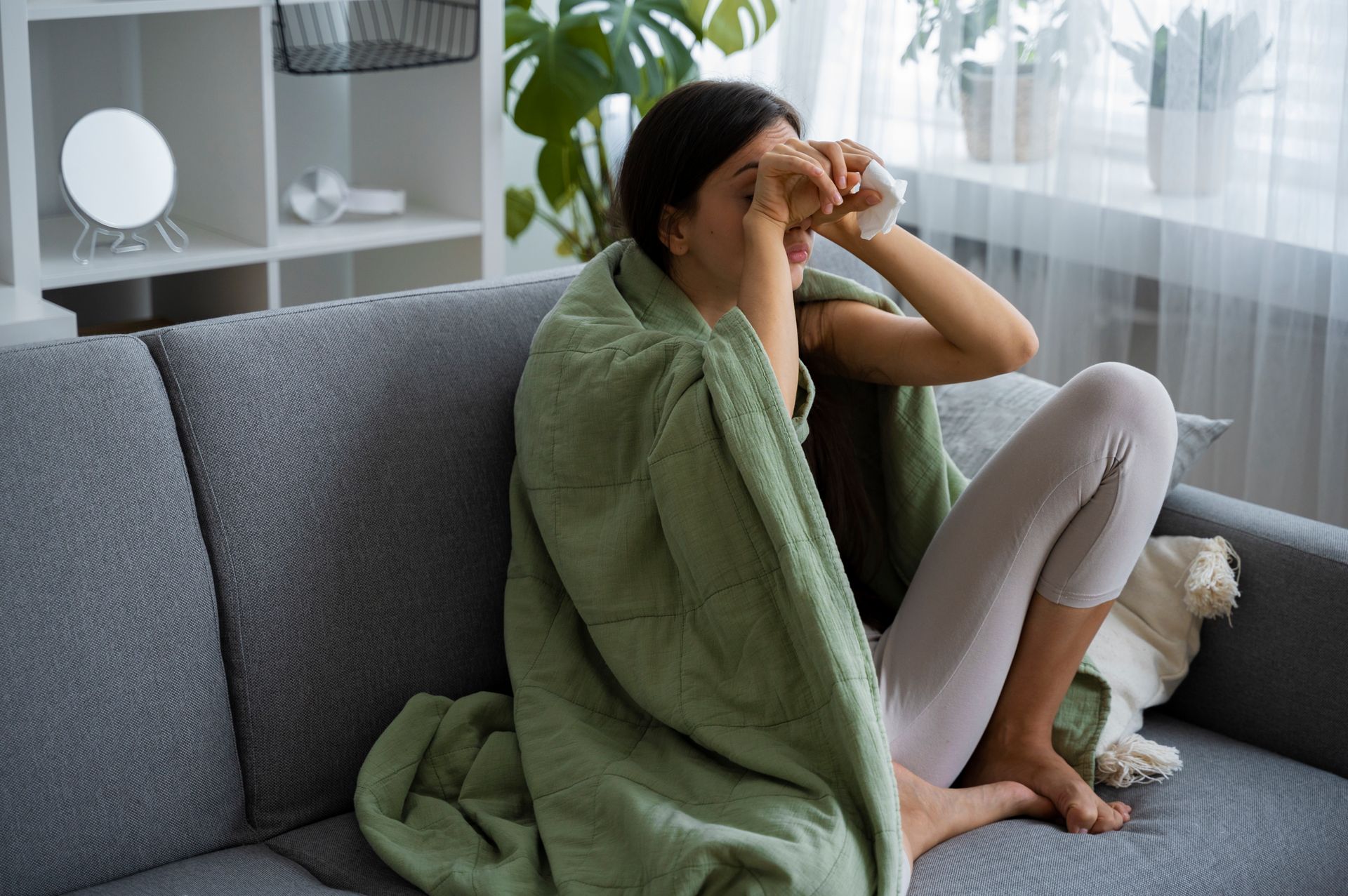 Woman on a couch covered in a green blanket, holding tissues near her eyes.