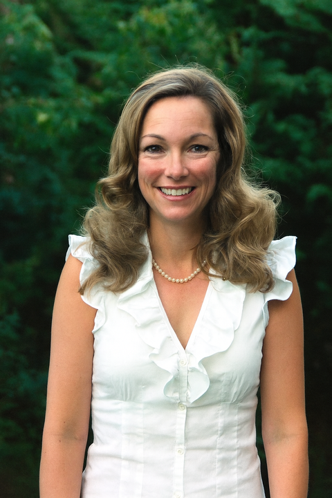 Woman with wavy blonde hair smiles, wearing white top and pearl necklace.
