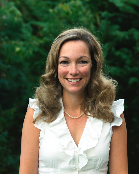 Woman with wavy blonde hair smiles, wearing white top and pearl necklace.