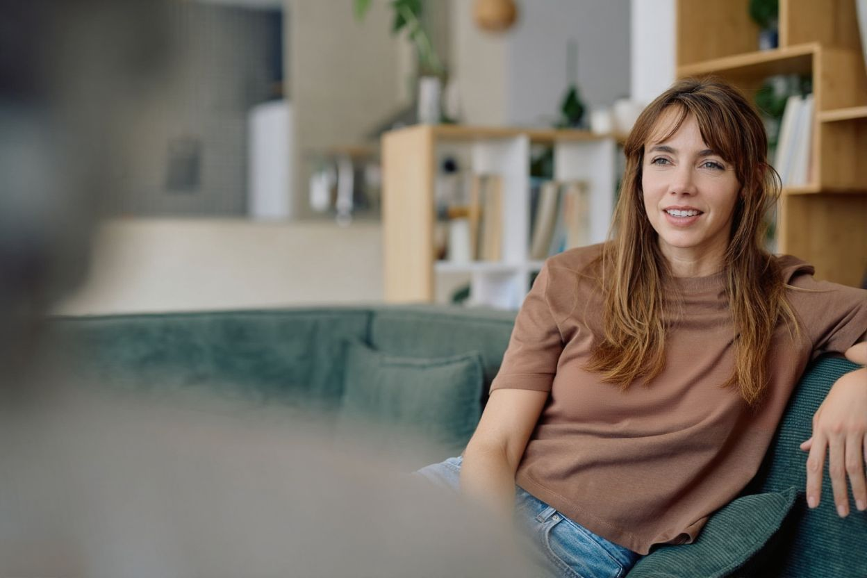 Woman in brown shirt on a teal couch, talking. Indoor setting with bookcase.