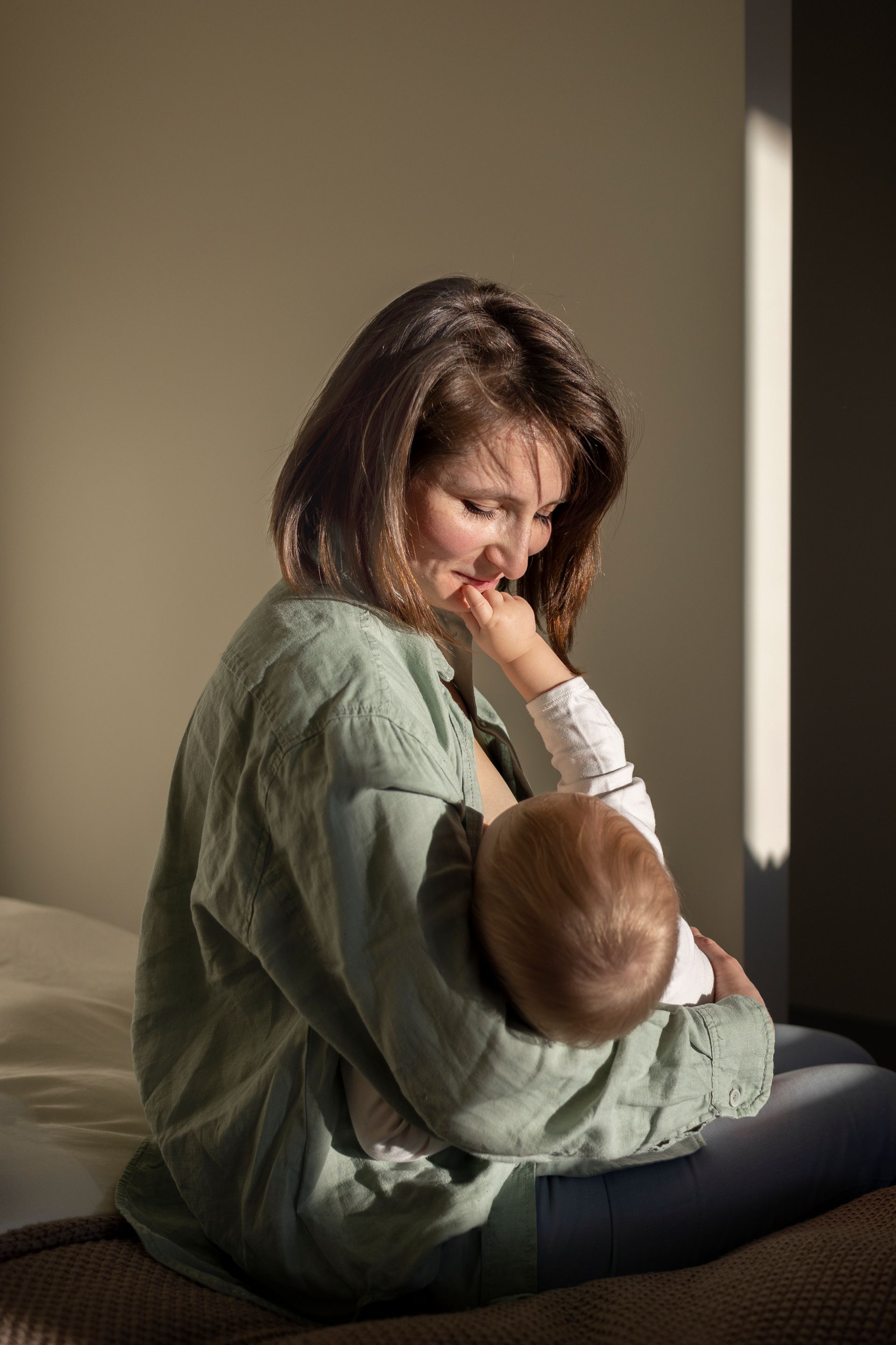 Woman breastfeeding a baby, sitting indoors in sunlight, wearing a green robe, looking down with a smile.