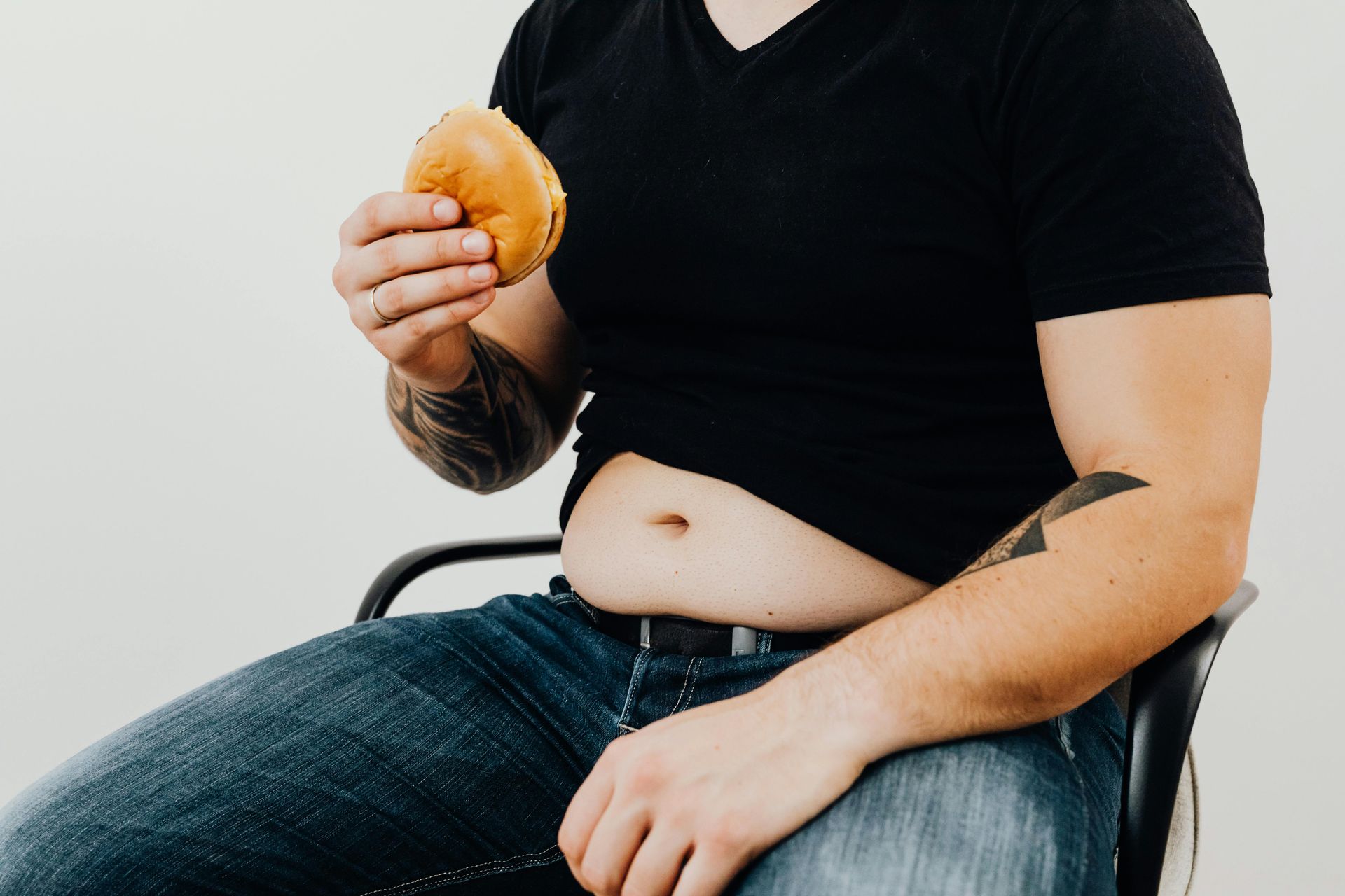 Person in black shirt and jeans holding a bun, seated; visible belly and arm tattoos.
