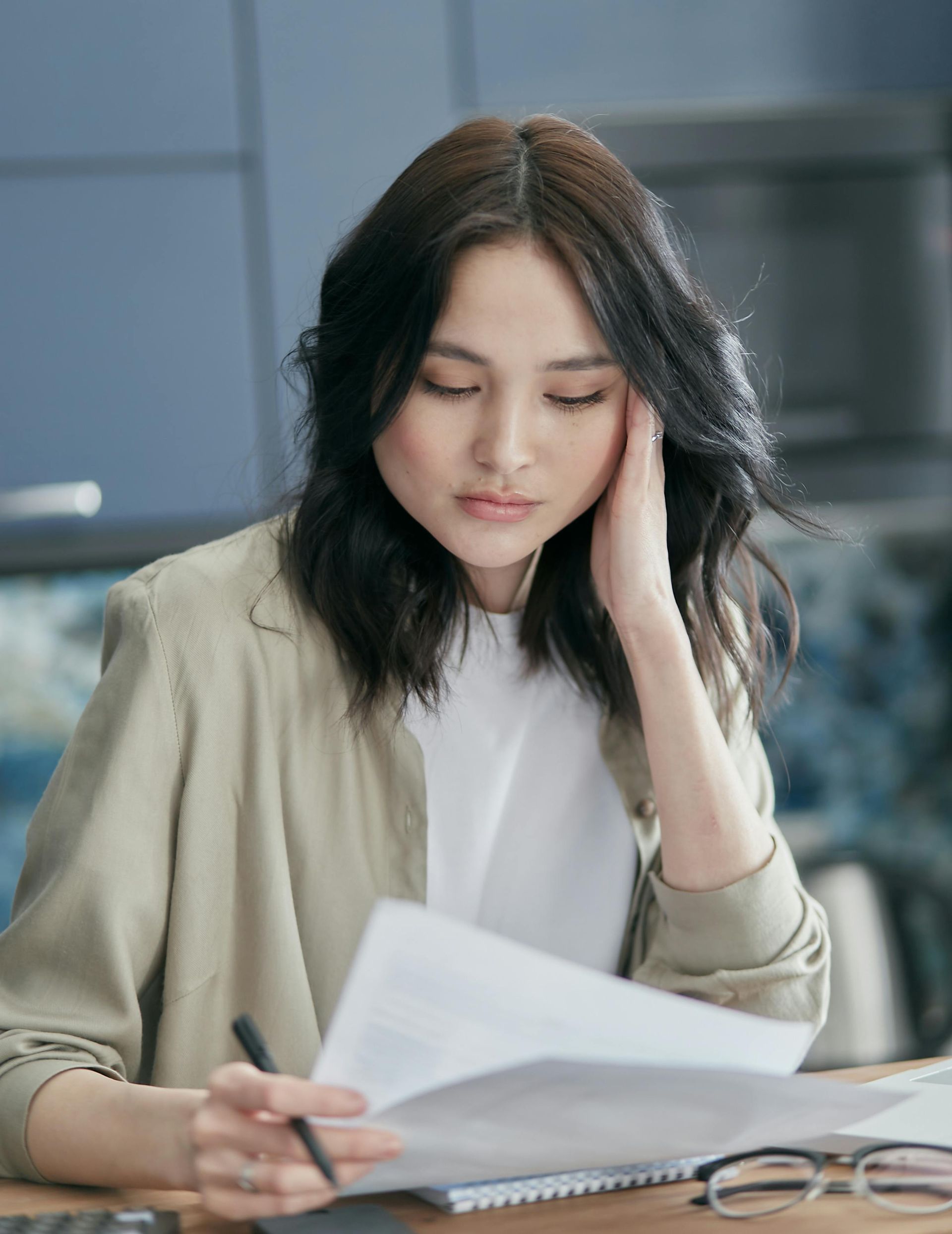 Woman in light jacket, looking at papers, holding her head, and pen in hand.