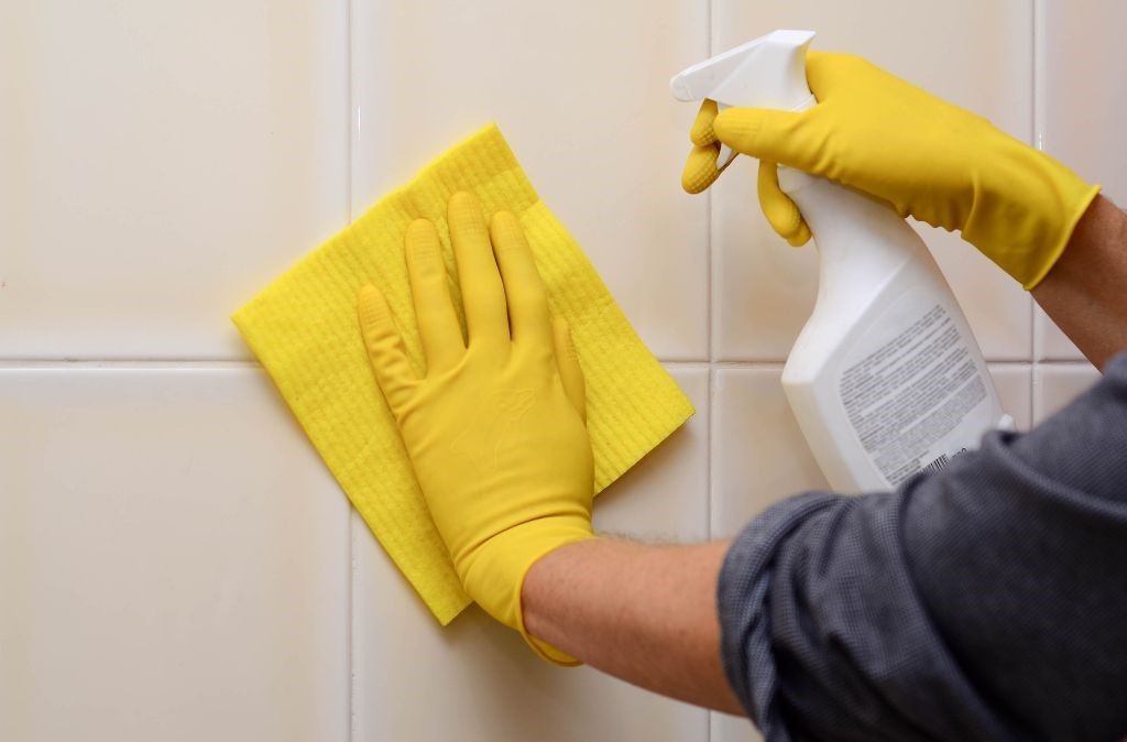 A person wearing yellow gloves is cleaning a tile wall with a cloth and spray bottle.