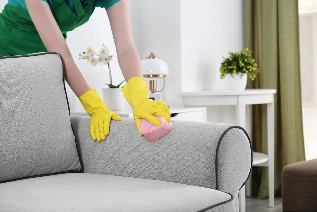 A woman in yellow gloves is cleaning a couch with a sponge.
