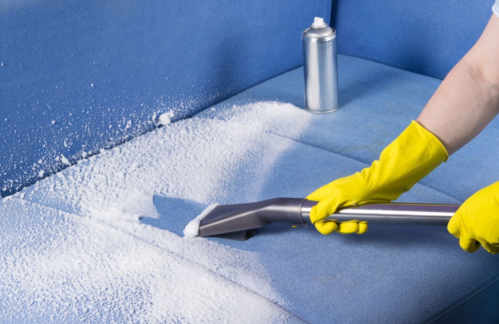 A person wearing yellow gloves is cleaning a blue couch with a vacuum cleaner.