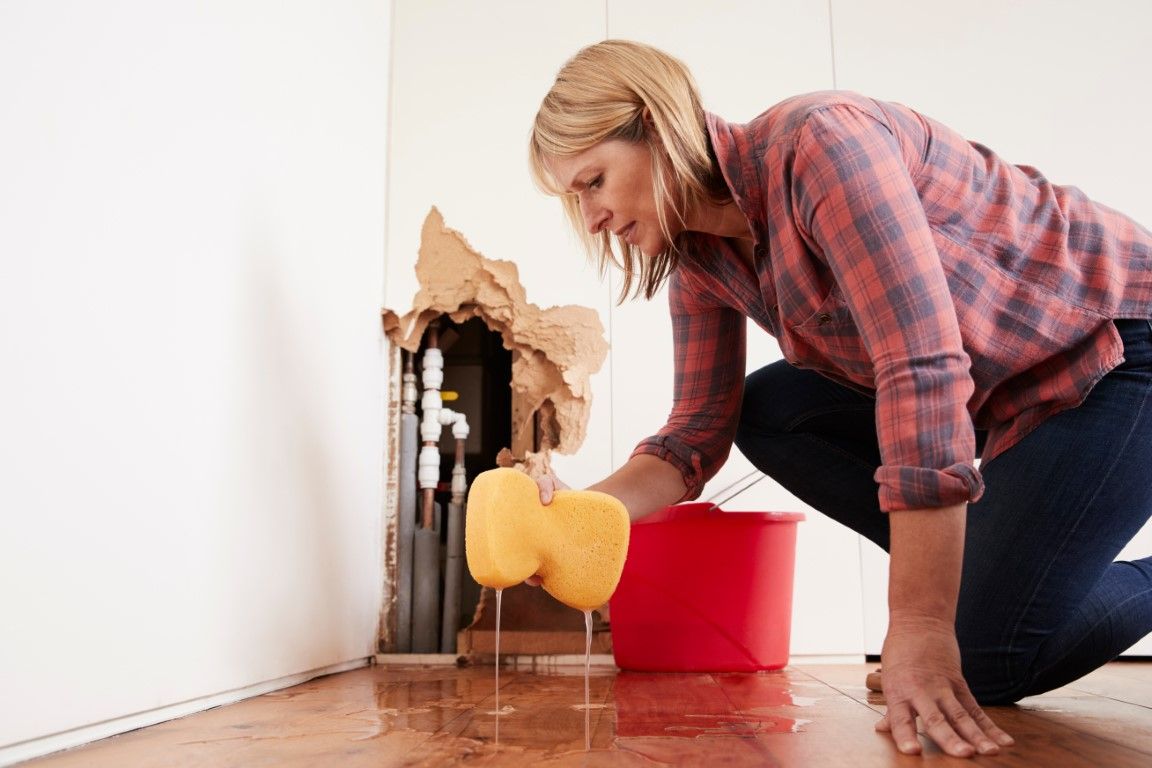 Woman mopping water from floor, next to burst pipe in wall. Red bucket.